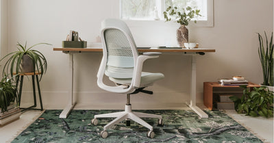 A home office setup features a Breck chair, a desk, and a rug. The desk has office supplies, a potted plant, and a notebook. The room includes several green plants and a window.