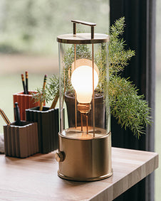 A modern, cylindrical Tala table lamp with a clear glass body and brass-finished base and top is lit on a wooden desk. Behind the lamp are a set of geometric pencil holders, a small potted plant, and a window.
